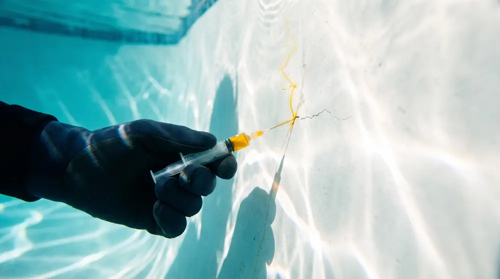 A gloved hand releases a thin line of yellow dye from a syringe beside a hairline crack in a white plaster pool wall, and the dye is drawn into the crack.